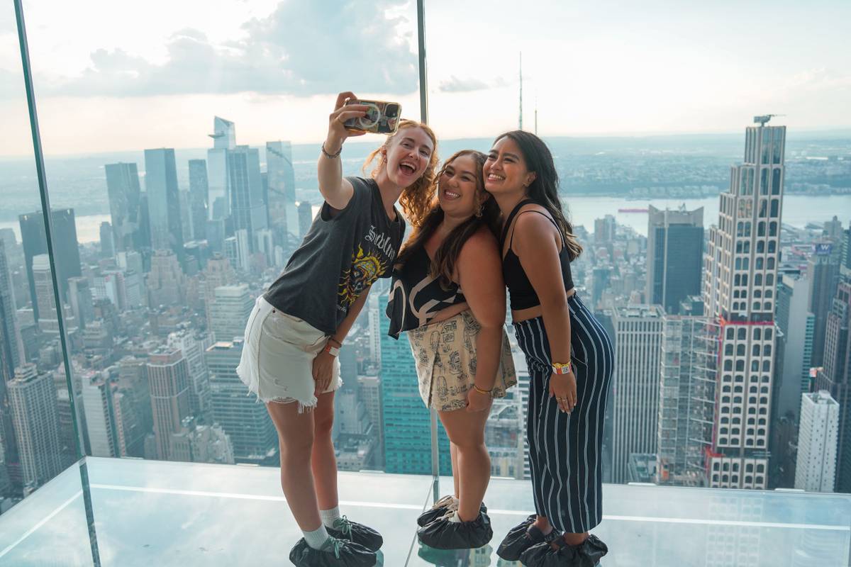 Visitors taking a selfie on the glass skydeck at Summit One Vanderbilt with the New York City skyline in the background.