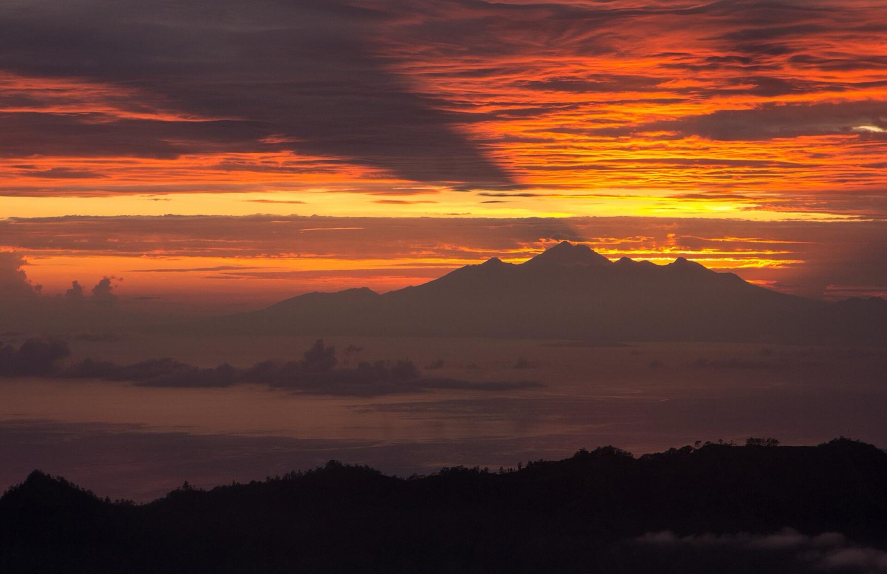 A red and orange dawn sky over the silhouette of Mount Rinjani on Lombok, seen across the sea from the summit of Mount Batur