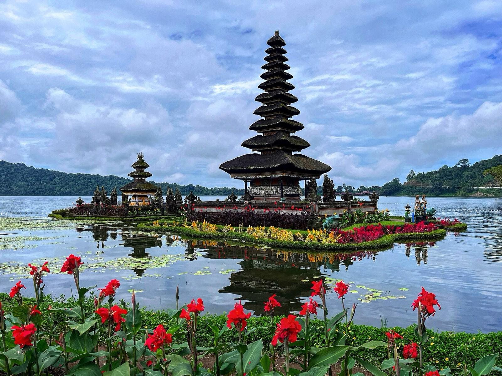 The tiered thatched shrine of Pura Ulun Danu Beratan standing on Lake Beratan, with red flowers in the foreground and forested hills behind