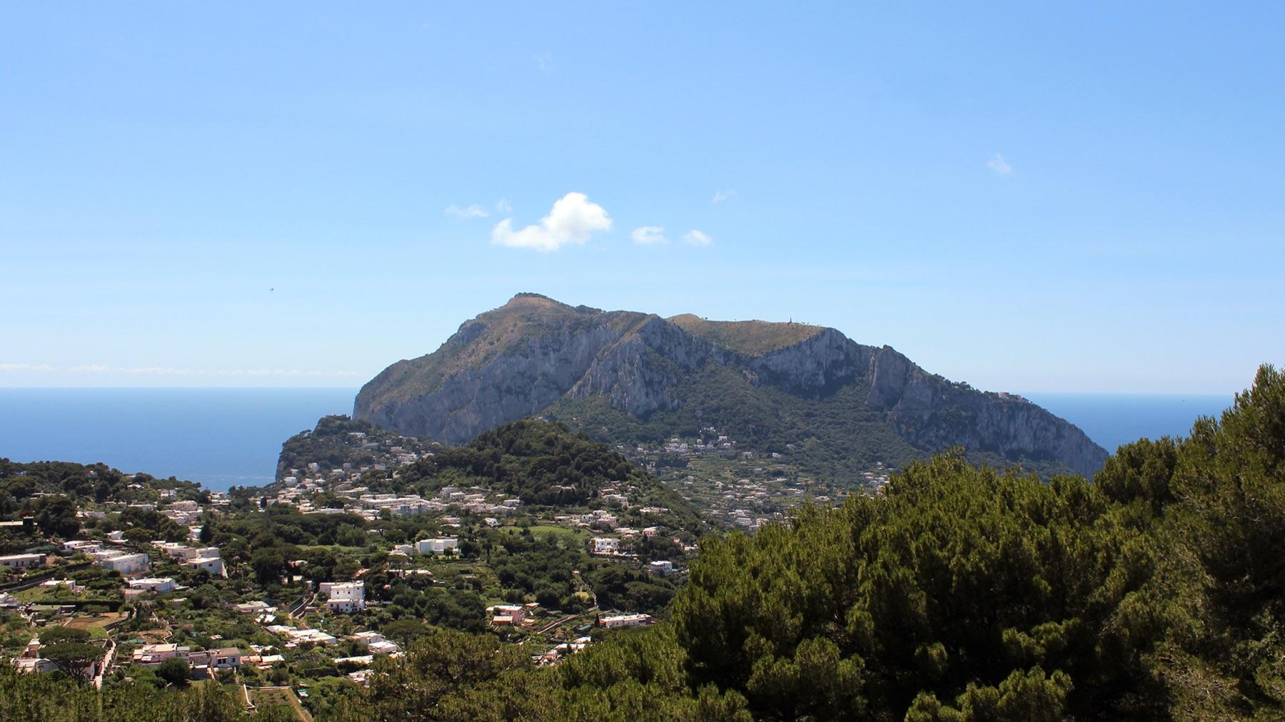 Monte Solaro rising above the white houses and pine trees of Capri under a blue sky, with the sea on both sides