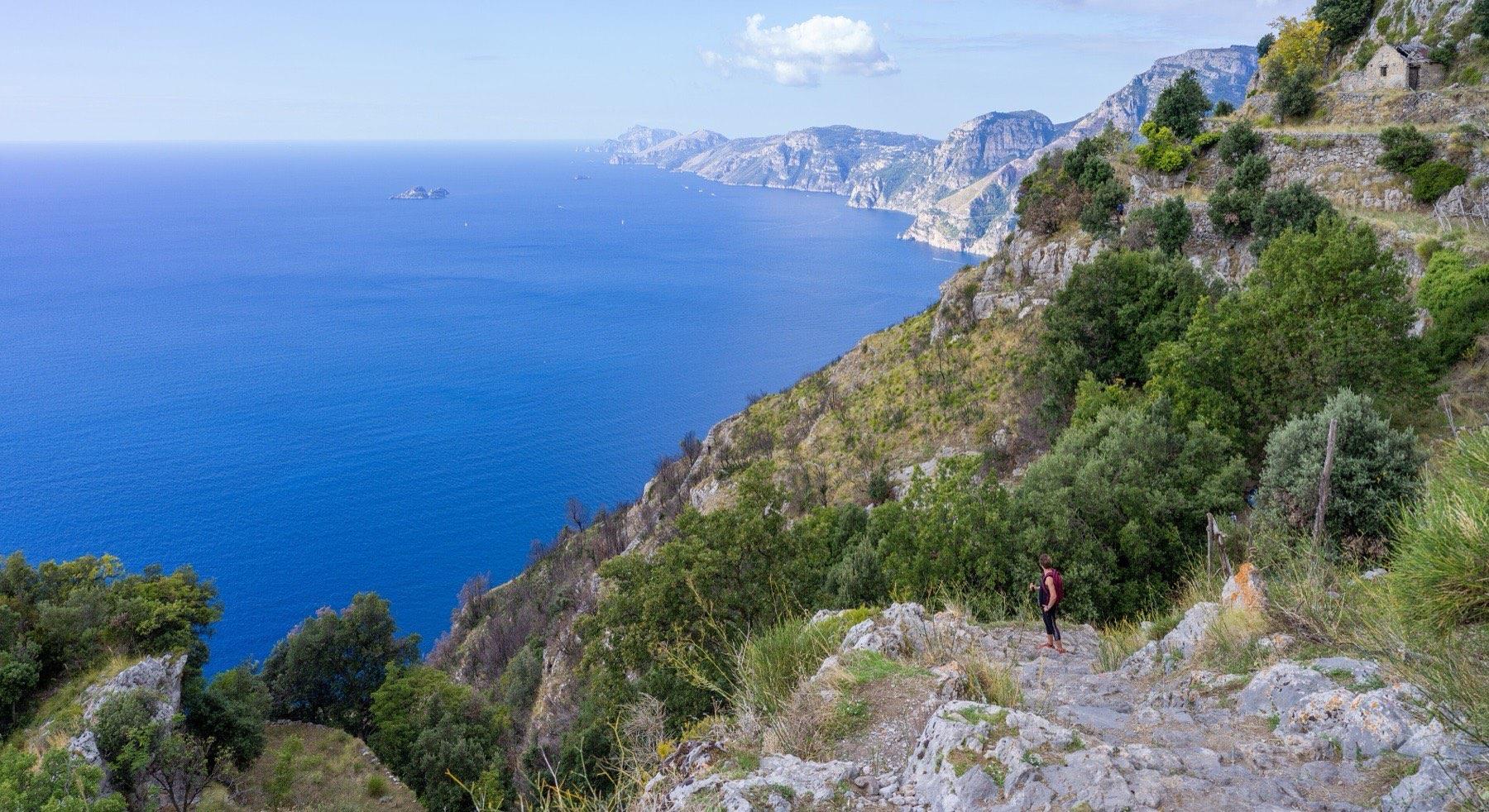 A walker on the stony Path of the Gods high above the blue sea, with the cliffs of the Amalfi Coast and the Li Galli islets in the distance