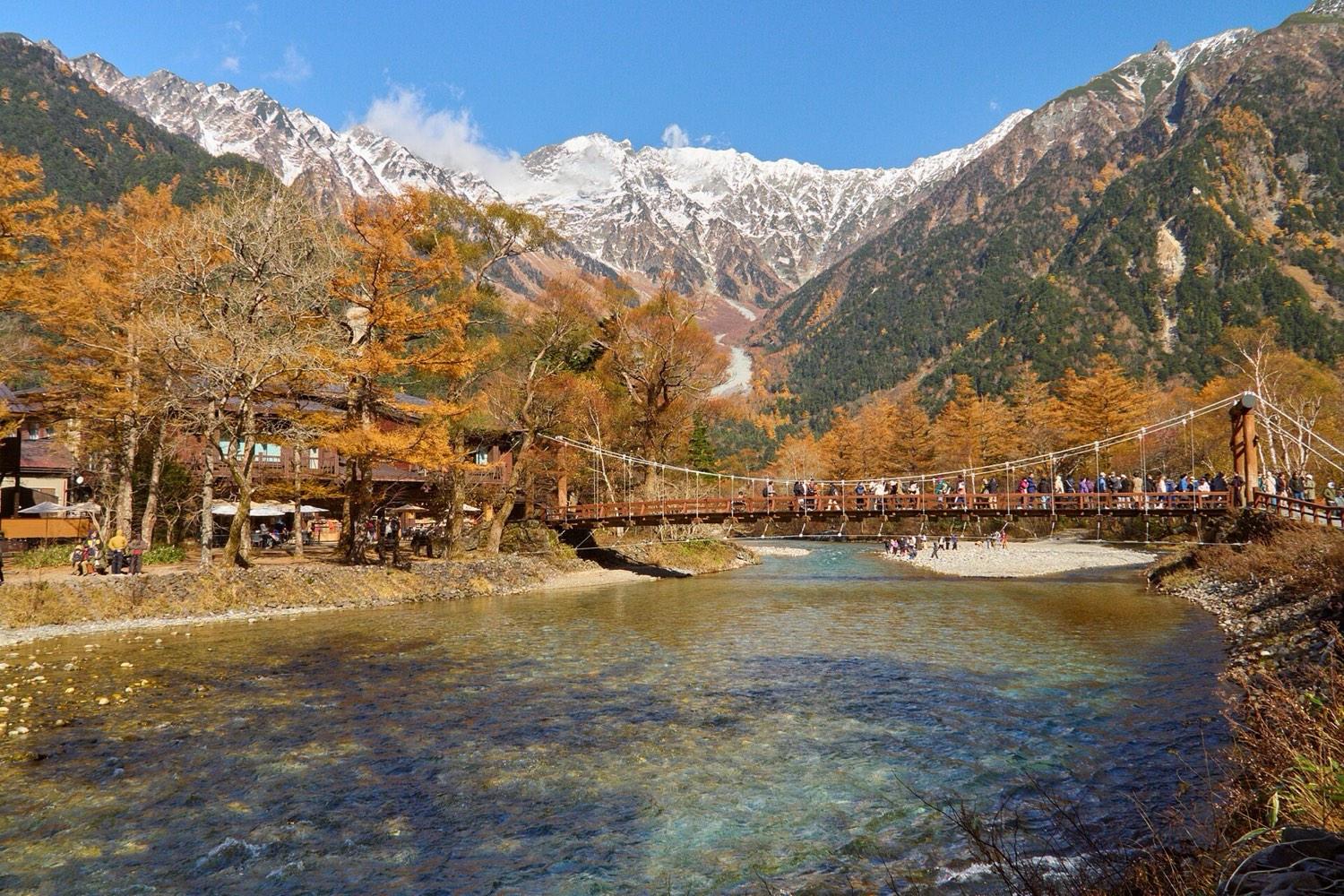 Kappa Bridge over the clear Azusa River in Kamikochi, with autumn larches and snowy Hotaka peaks behind