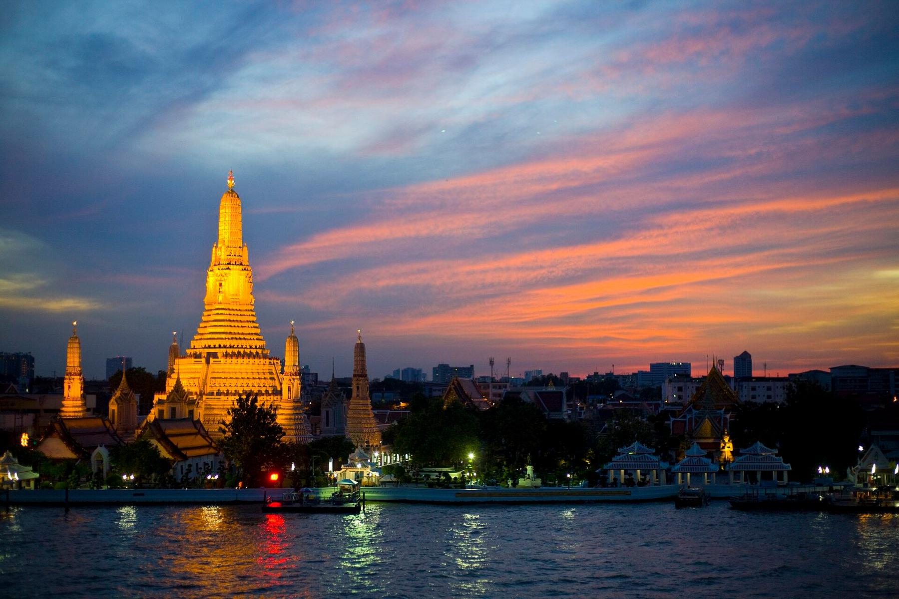Wat Arun lit gold at dusk on the bank of the Chao Phraya River under a pink and violet sky