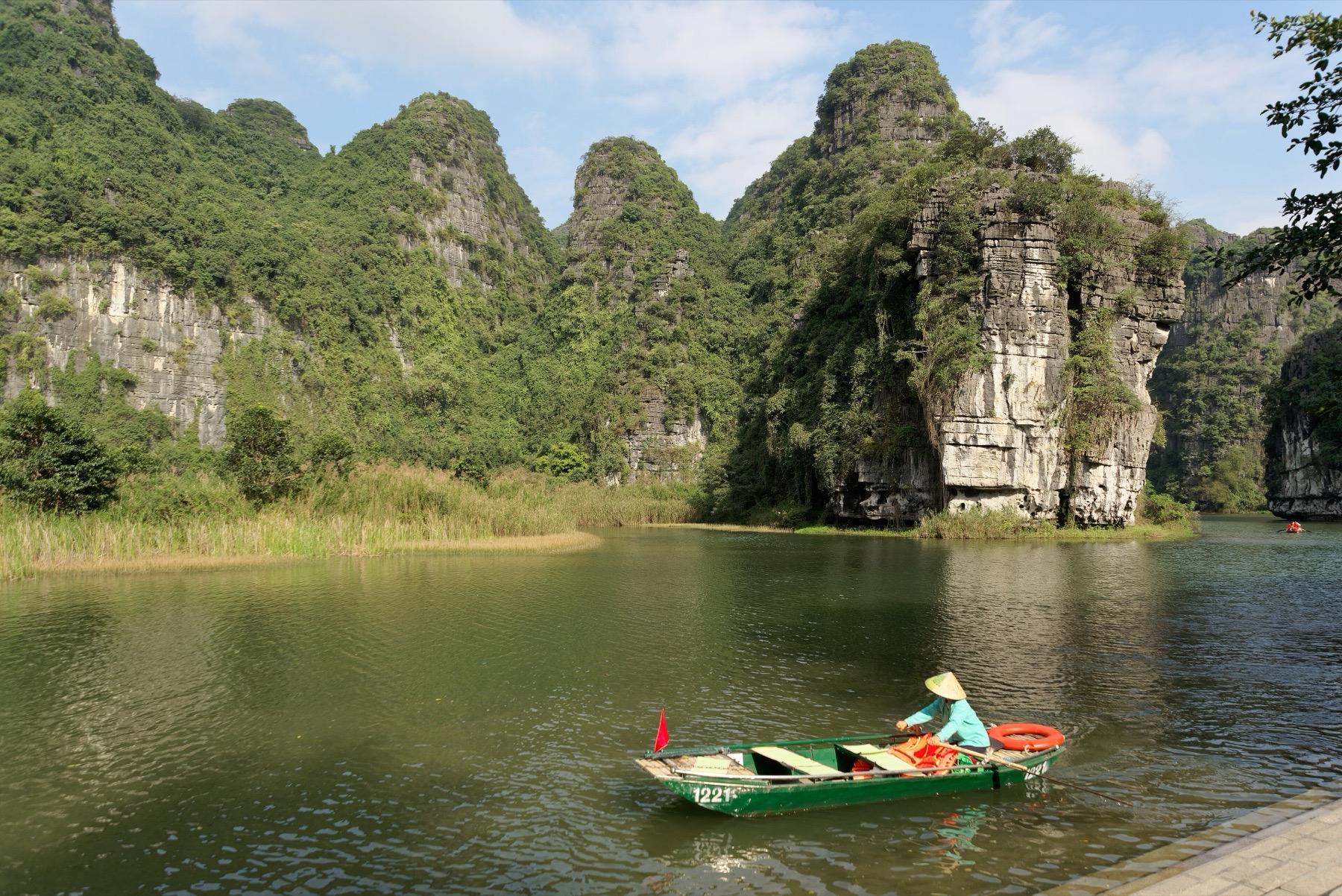 A rower in a conical hat in a small green boat on the river below forested limestone cliffs at Trang An