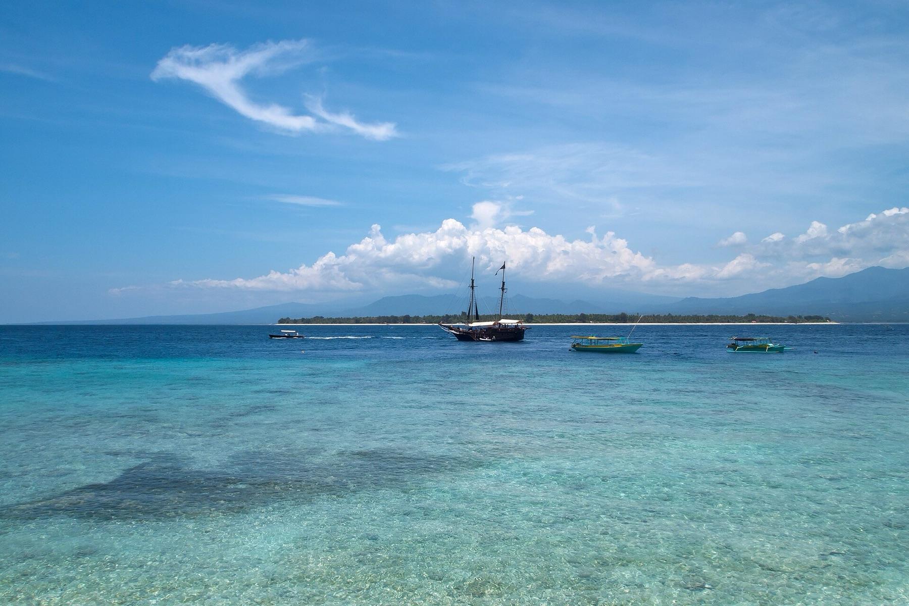 Clear turquoise water and moored boats off Gili Meno, with the low green line of Gili Air and the mountains of Lombok behind