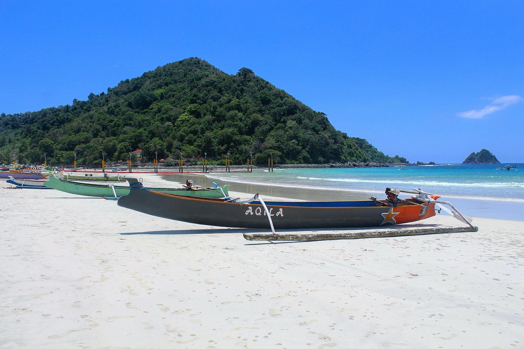 Painted wooden outrigger fishing boats pulled up on the white sand of Selong Belanak, with a green hill and blue water behind