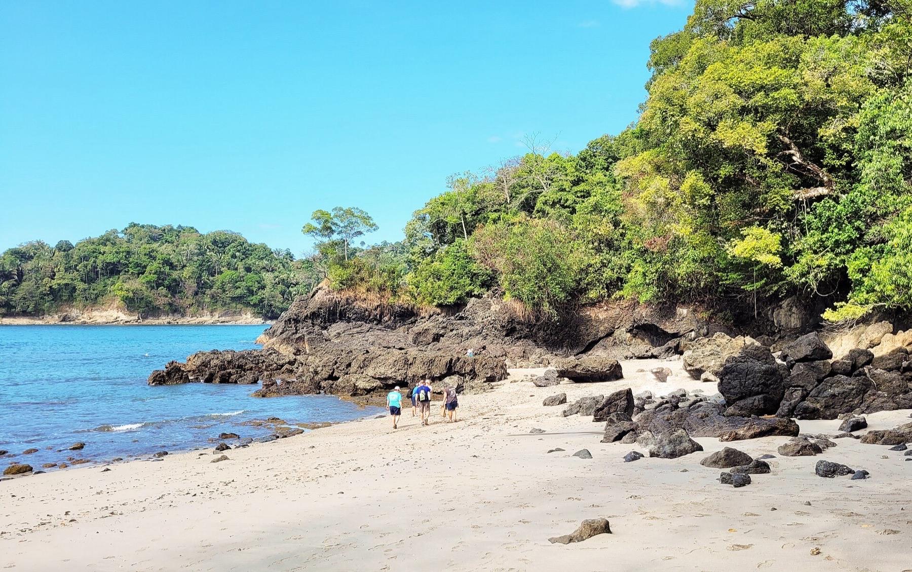 Walkers on a pale sand beach in Manuel Antonio National Park, with forested headlands and a calm blue sea