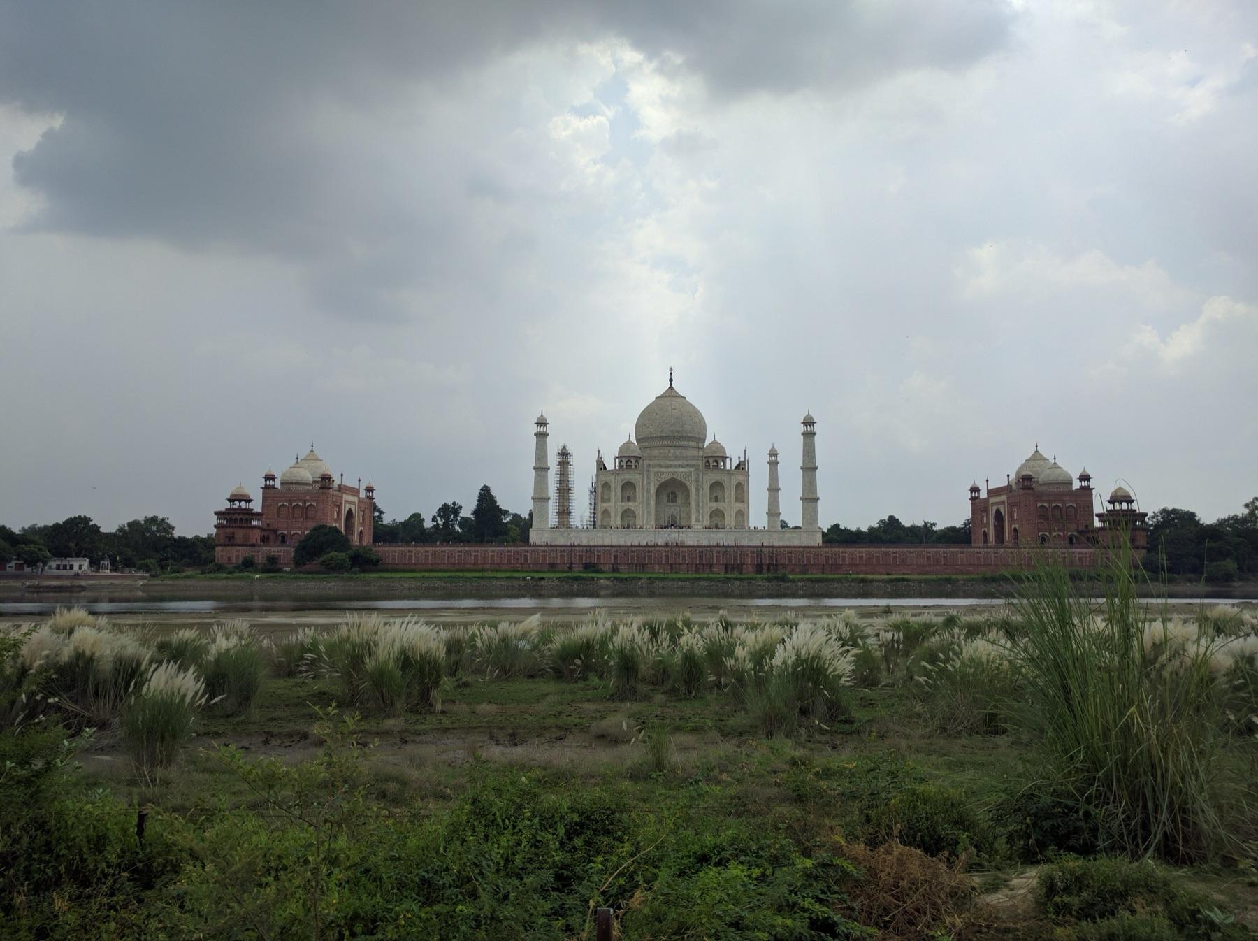 The Taj Mahal seen across the Yamuna River from the grasses of Mehtab Bagh under a cloudy sky