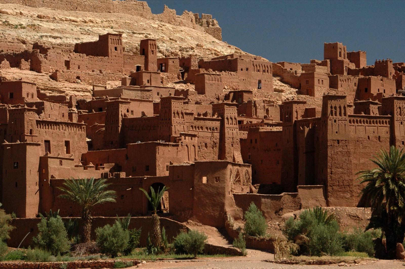 The earth-built towers and walls of the ksar of Ait Benhaddou rising up a hillside behind palm trees