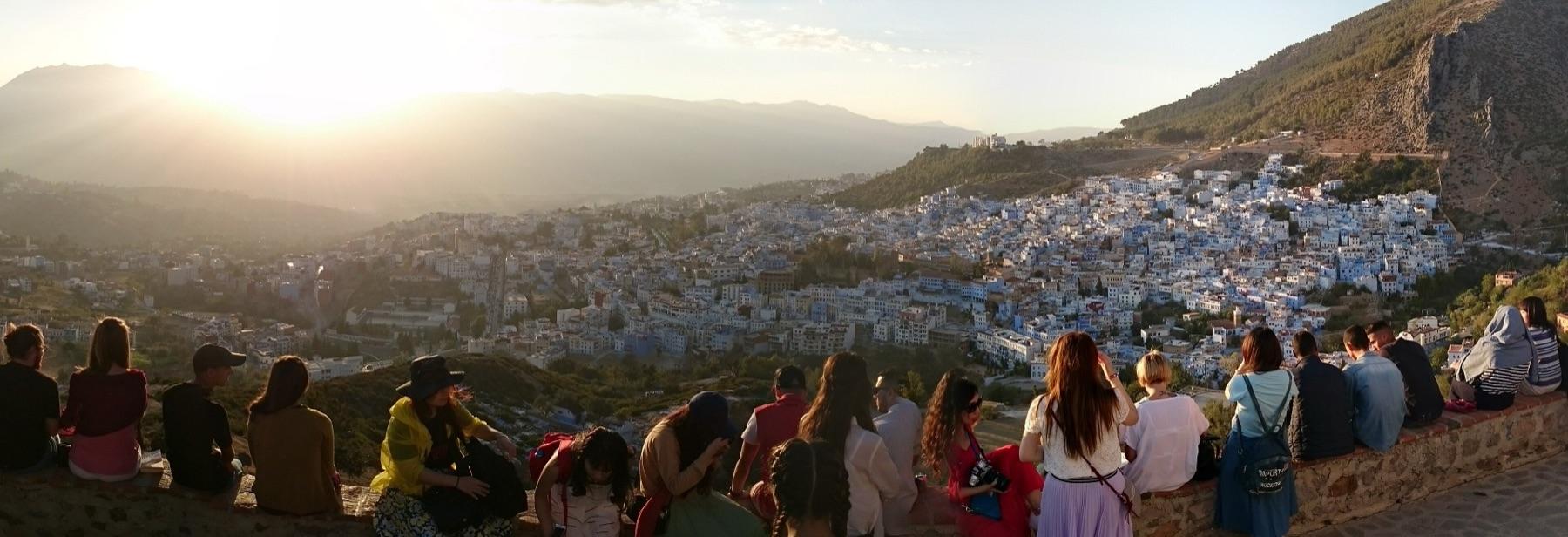 Visitors sit on the wall by the Spanish Mosque above Chefchaouen as the sun goes down over the blue town and the Rif Mountains