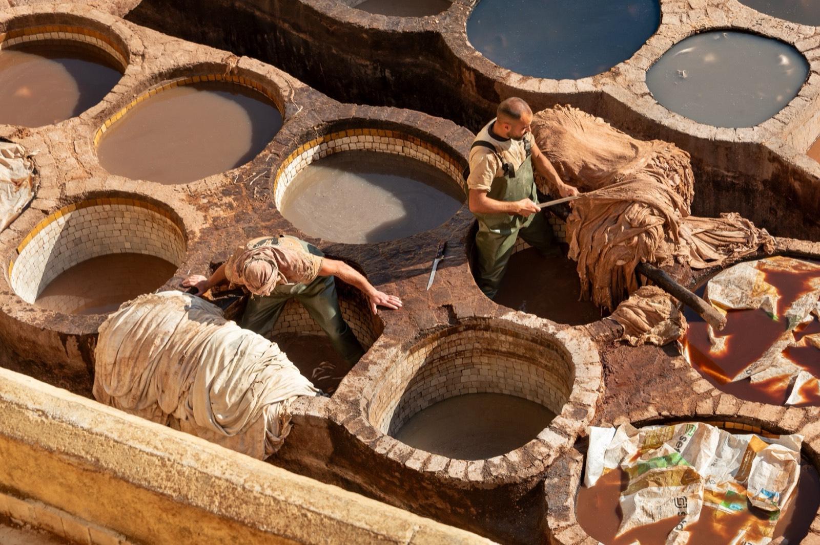 Two tanners work among the round stone dye pits of the Chouara tannery in Fes, seen from a terrace above