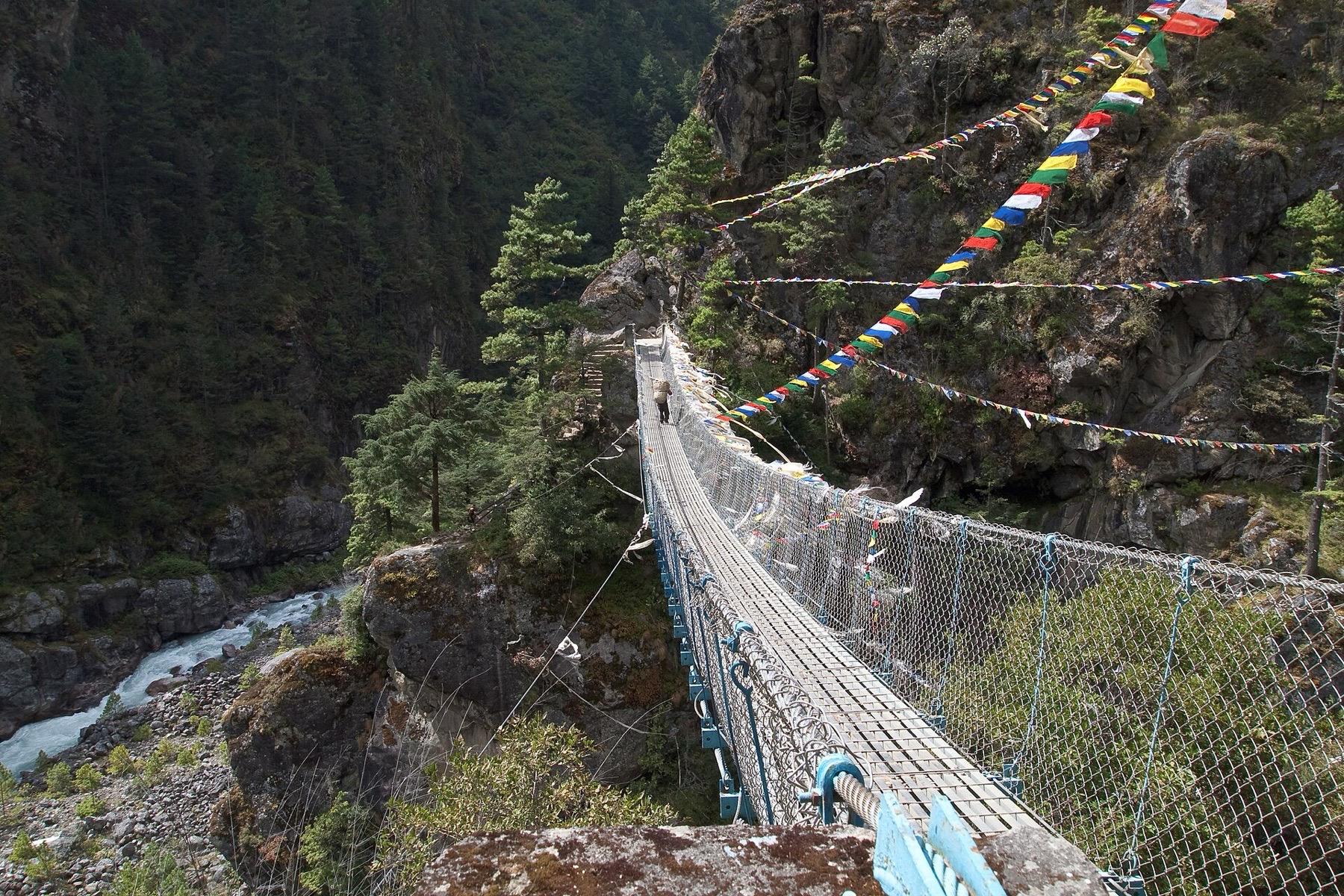 A suspension footbridge hung with prayer flags crosses a forested gorge above the Dudh Koshi river