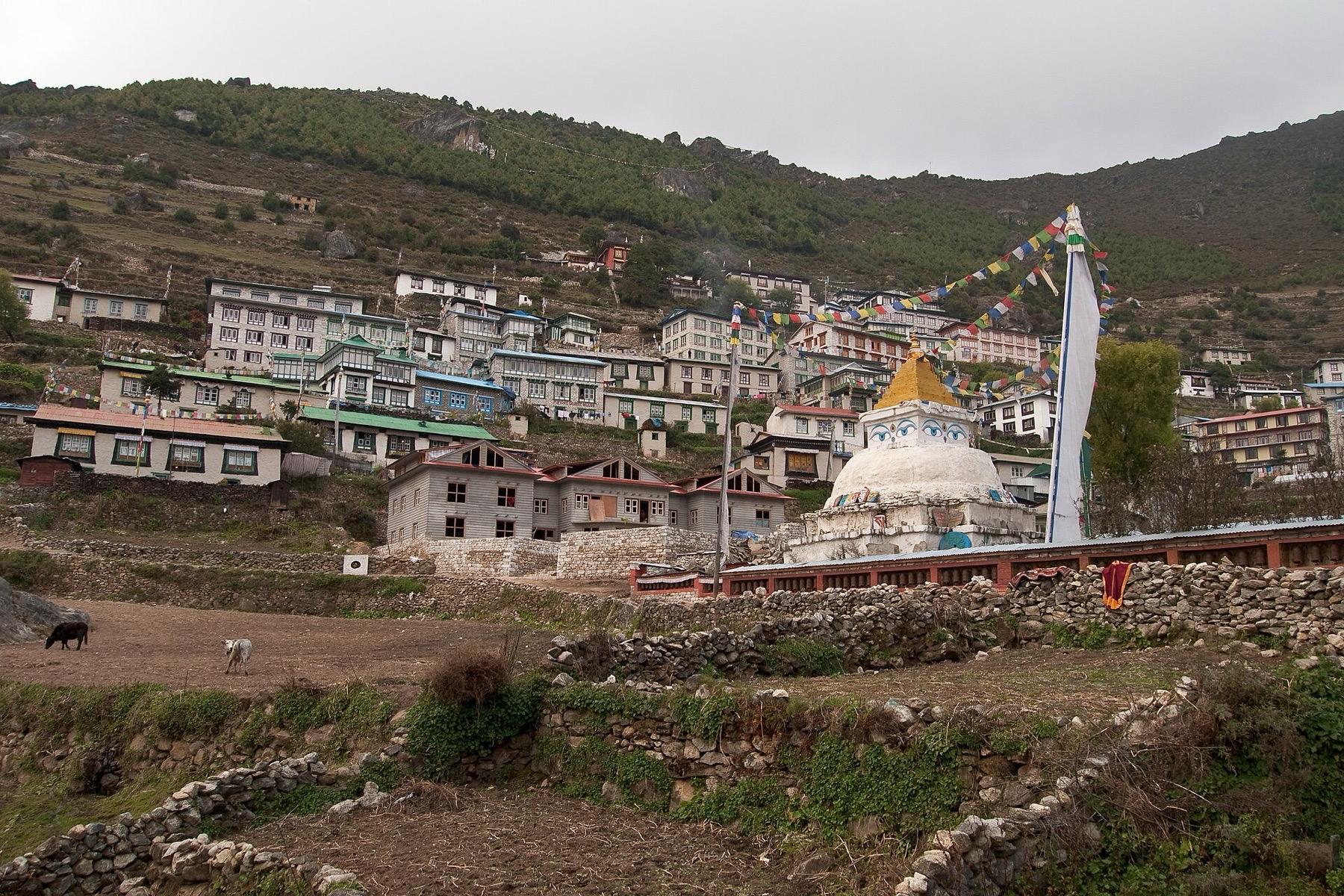 A white stupa with prayer flags below the terraced stone and tin-roofed houses of Namche Bazaar