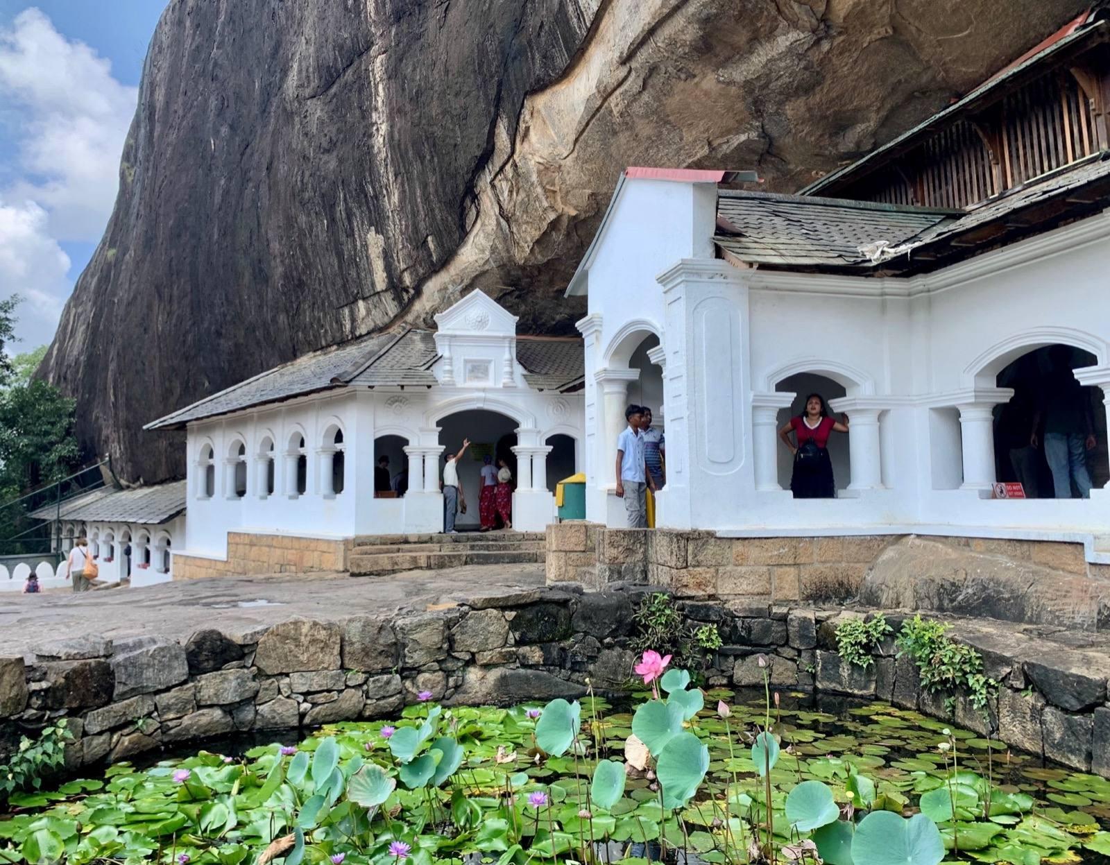 The white arched entrance of the Dambulla cave temple under its overhanging rock, with a lotus pond in front