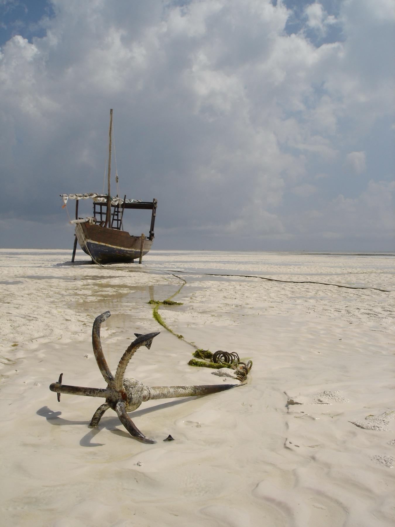 An old anchor lies on the white sand at low tide on Zanzibar's east coast, with a wooden dhow beached behind