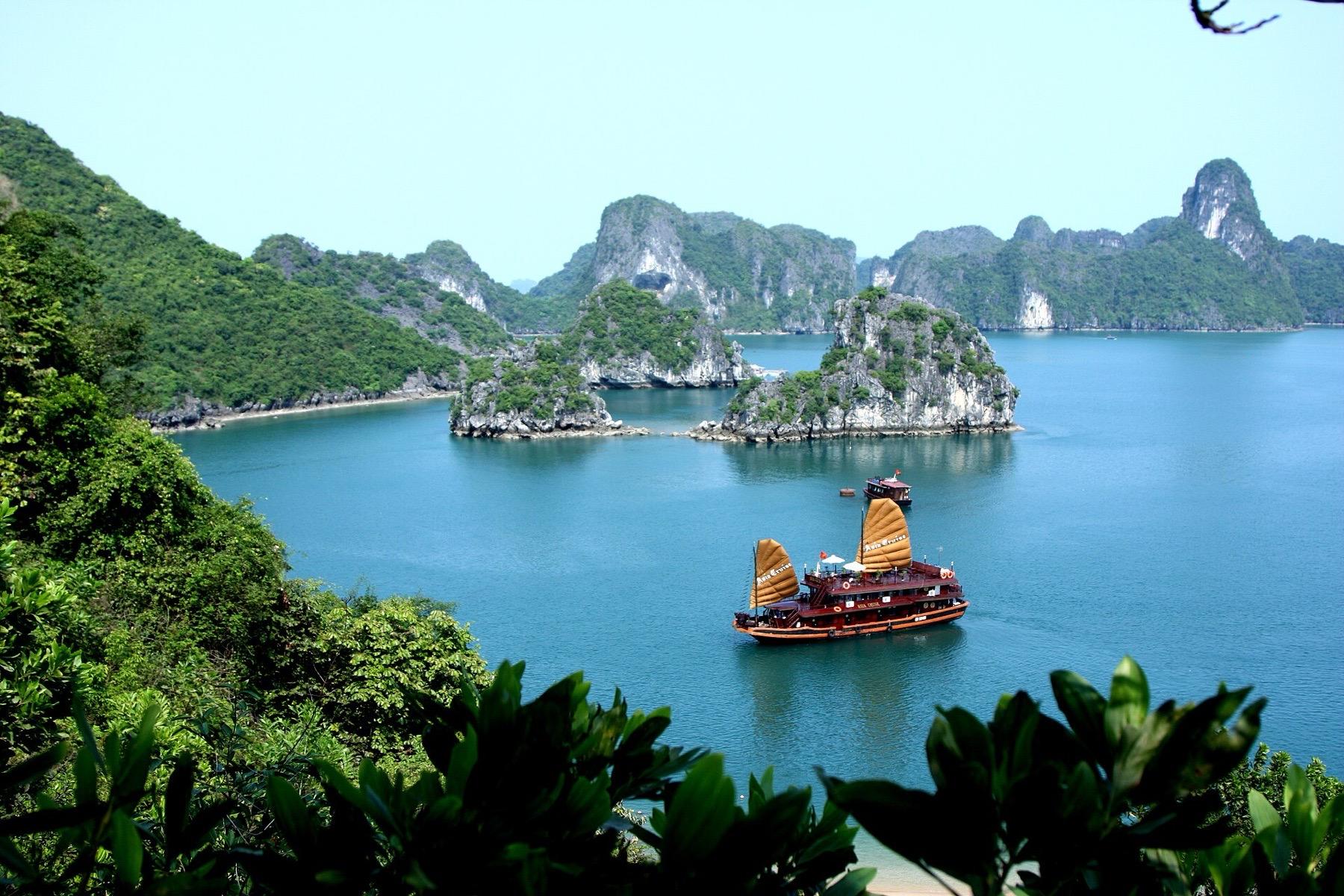 A wooden junk with tan sails anchored among forested limestone islands in Ha Long Bay