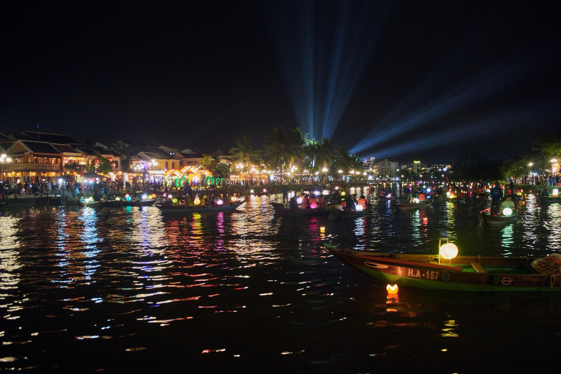 Small boats with lit lanterns on the Thu Bon River at night, with the lights of Hoi An Old Town reflected in the water